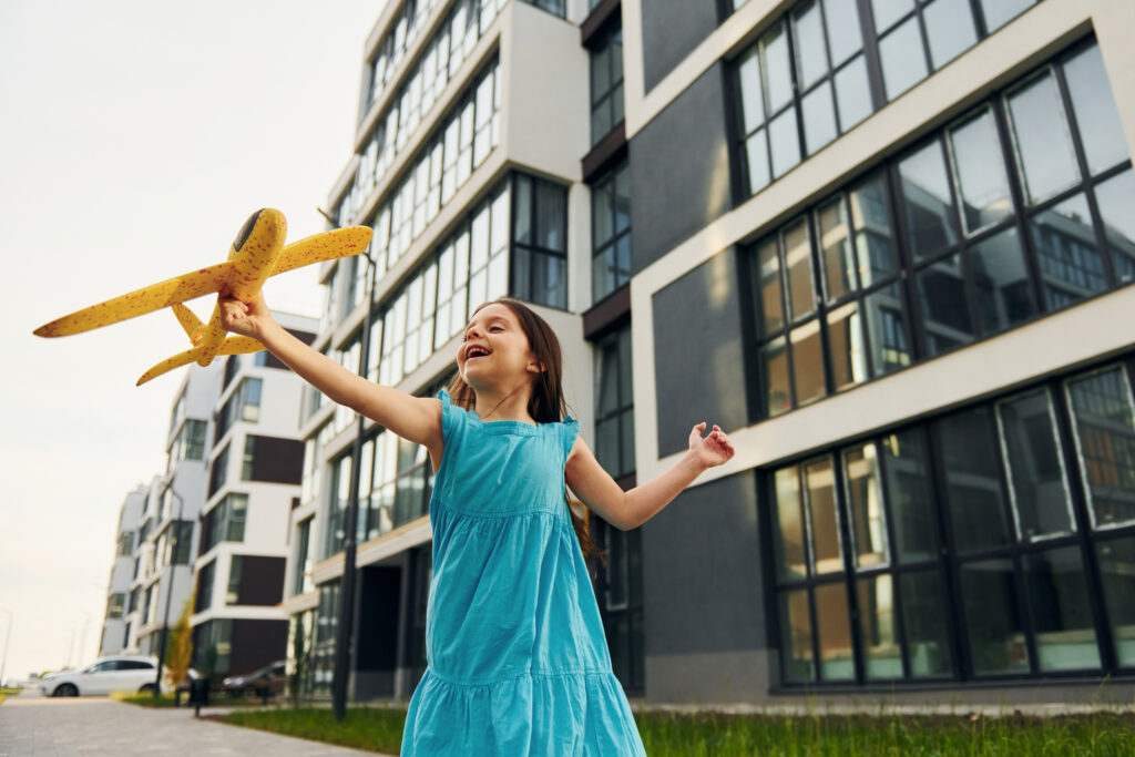 With toy plane. Cheerful little girl walking outdoors in the city in dress.
