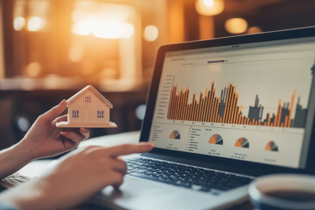 A person is holding a miniature house model while pointing at a vibrant graph displayed on a laptop screen, set in a well-lit indoor environment perfect for business analysis.